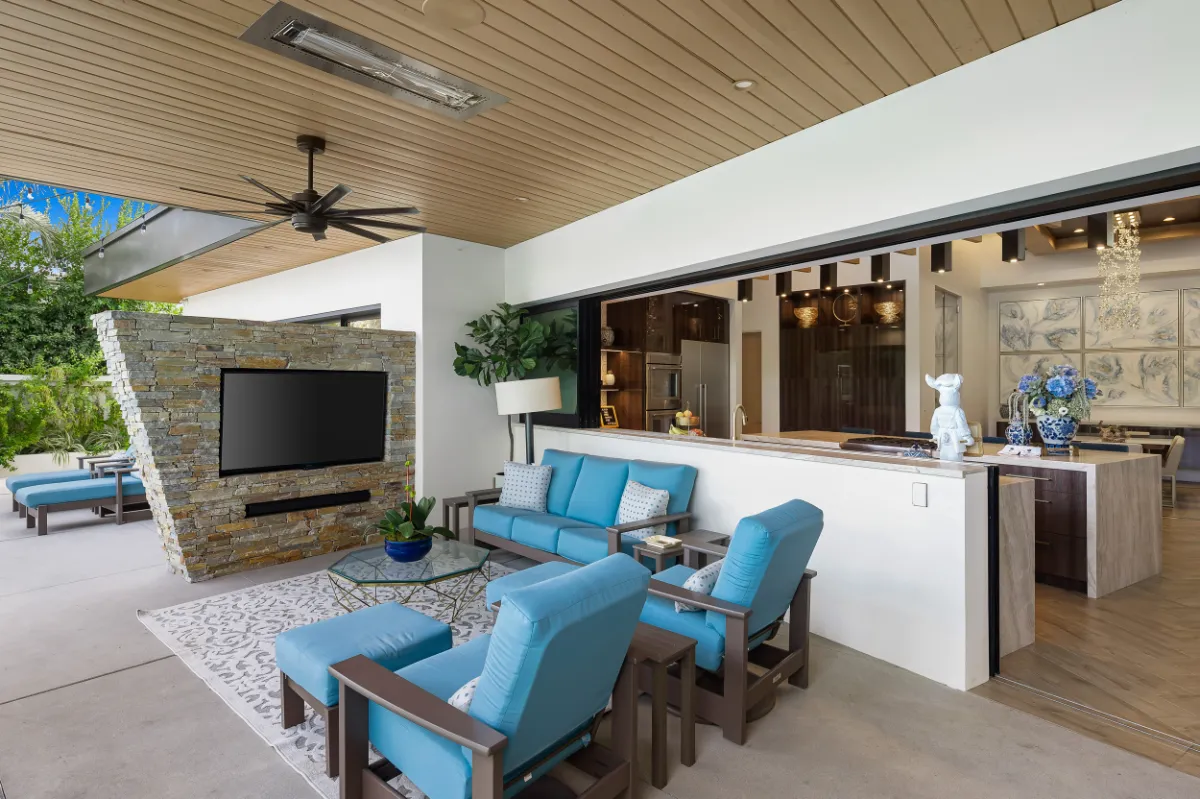 Covered outdoor room with a wood-slat ceiling, ceiling fans, blue lounge seating and a stone television wall