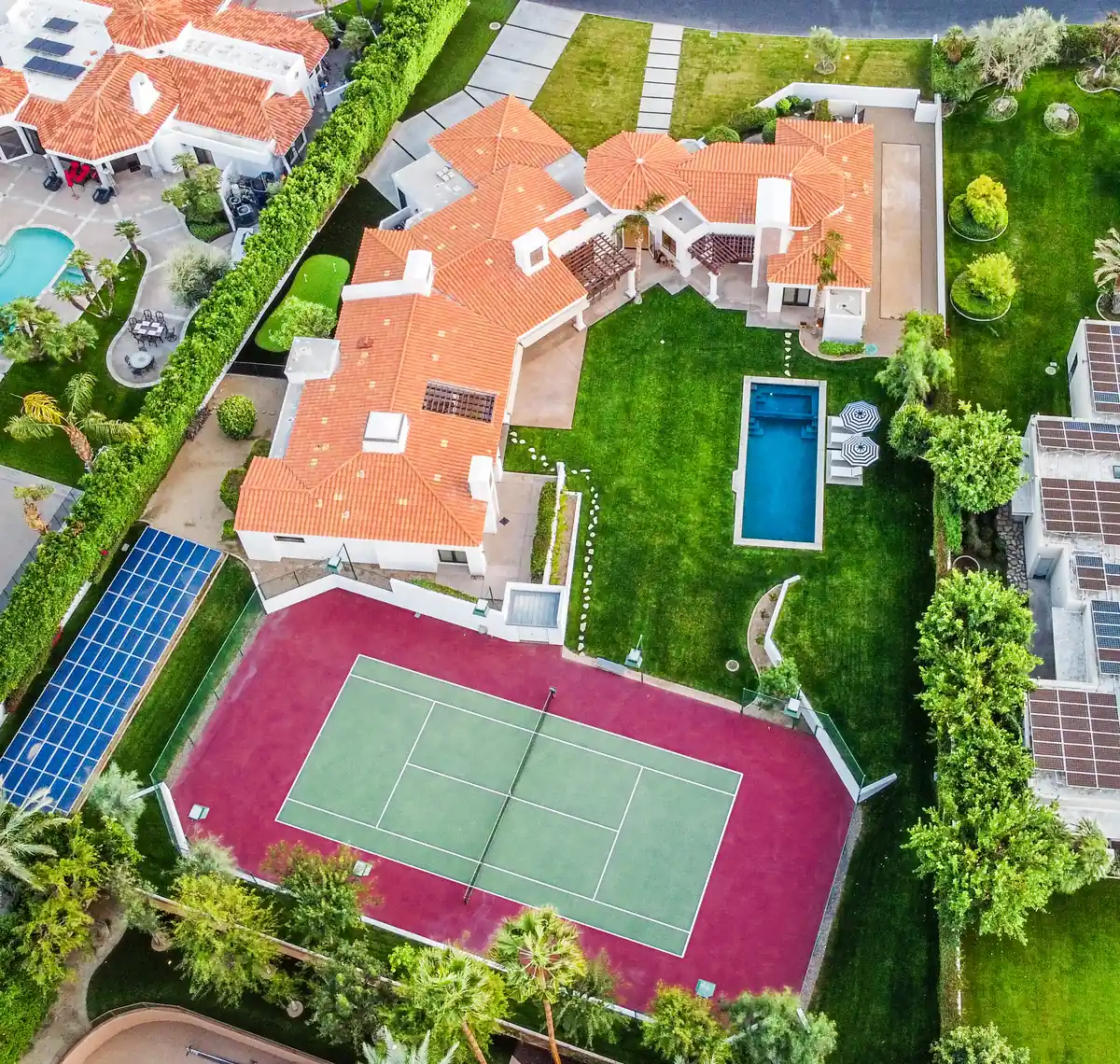 Aerial view of a walled estate with a tile roof, tennis court, pool and mature landscaping