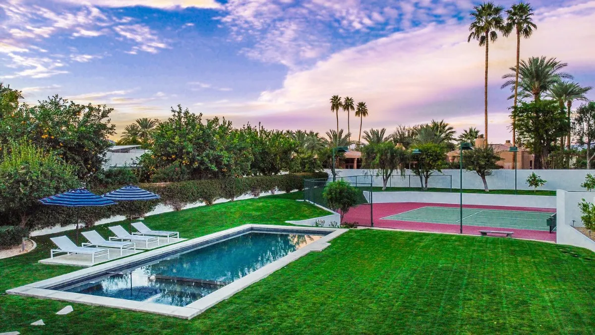 Lawn at dusk with a rectangular lap pool, loungers under blue umbrellas and a fenced tennis court