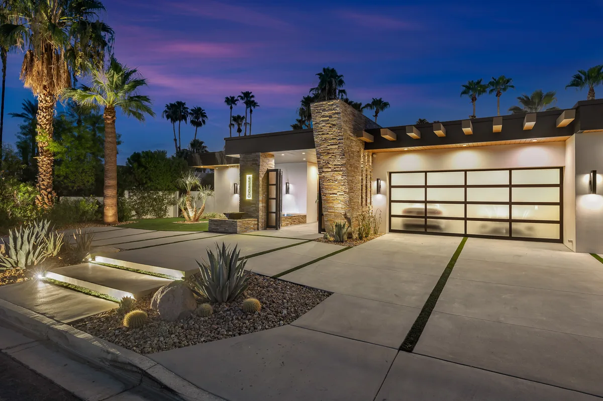 Front elevation at dusk with a glass garage door, stone column and a low fire feature in the desert planting