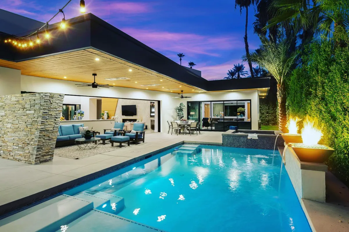 Backyard pool at dusk beside a covered patio with string lights and a lit fire bowl
